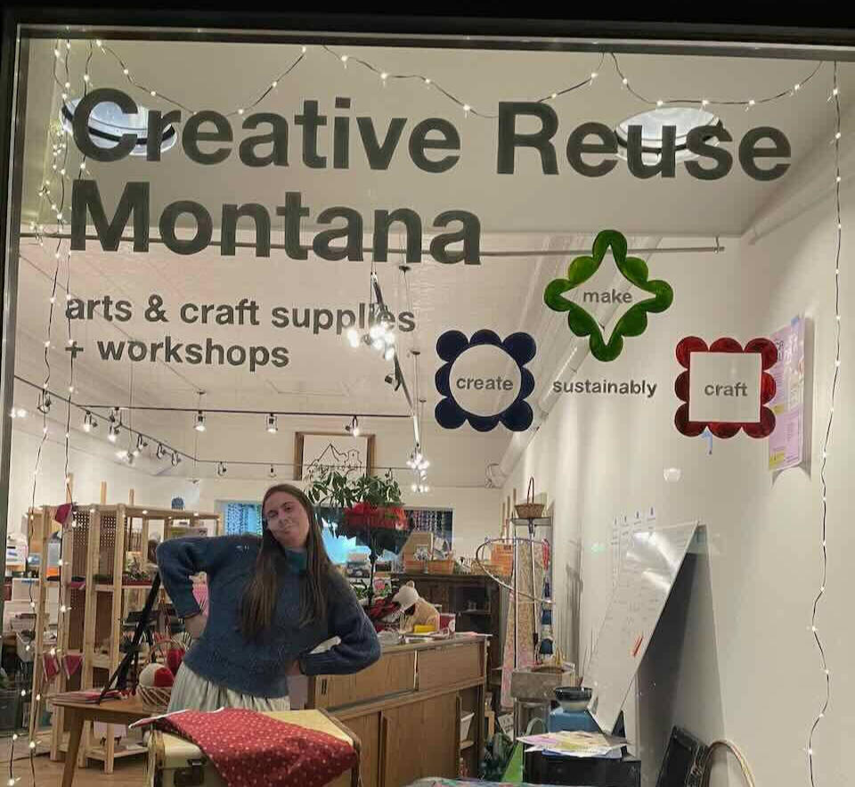 Image shows a store window with the words "Creative Reuse Montana" in bold text across the top and a view into a brightly lit store with art supplies on shelves and a friendly person with long brown hair and a blue sweater striking a pose